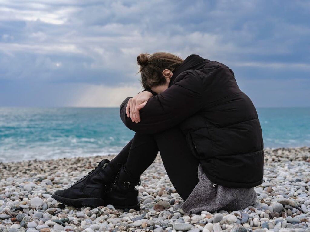 Mujer sentada cabizbaja en la playa, reflejando el sufrimiento mental tratado por tu psicóloga en Marbella.